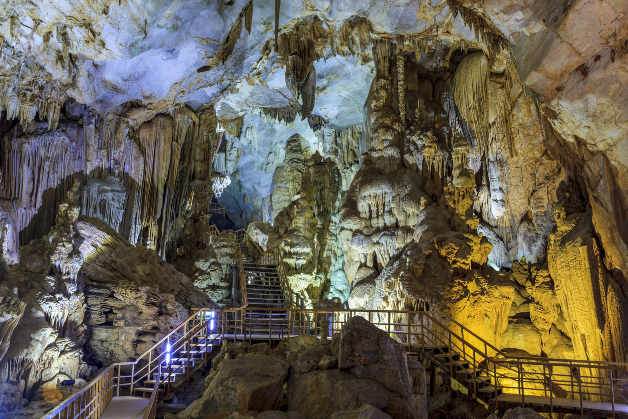 Beleuchtete Stalaktiten und Stalagmiten umgeben Holzstege in einer Höhle.