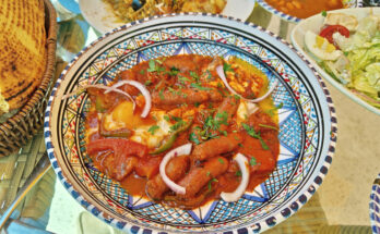 Bunt gemusterte Schale mit Ojja aus Tomatensauce, Merguez, pochiertem Ei, Zwiebelringen und Kräutern, daneben Brot und Beilagen.