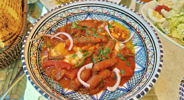 Bunt gemusterte Schale mit Ojja aus Tomatensauce, Merguez, pochiertem Ei, Zwiebelringen und Kräutern, daneben Brot und Beilagen.
