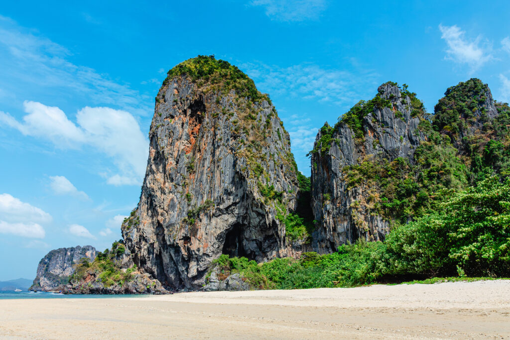 Nahaufnahme der Kalksteinwände am Phra Nang Beach mit leerem Strand, Vegetation und blauem Himmel.