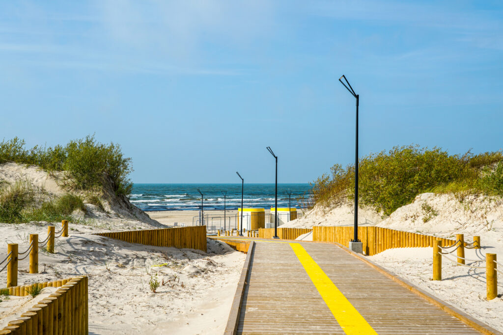 Barrierearmer Holzsteg mit gelber Linie führt durch Sanddünen zum Ostseestrand in Liepāja.