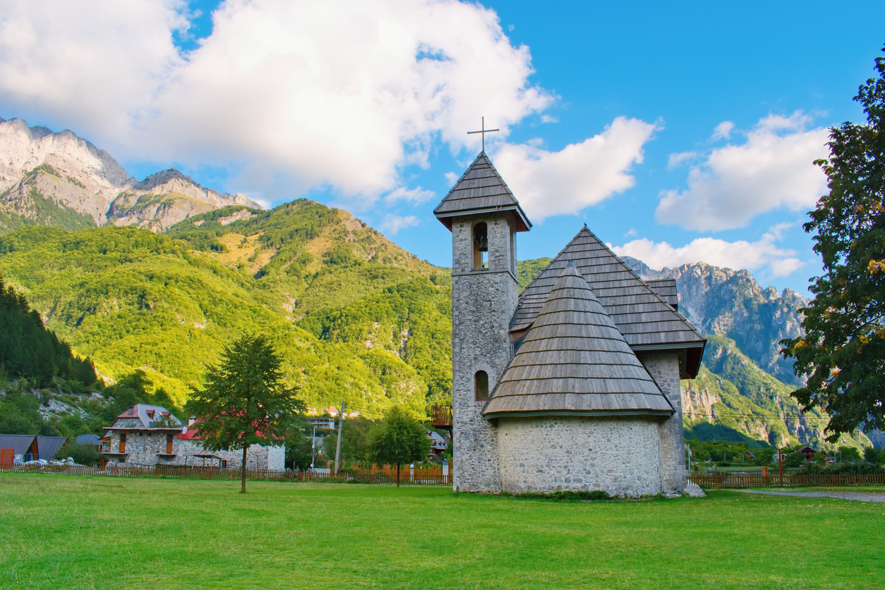 Eine kleine Kirche mit Schindeldach und Glockenturm steht auf einer Wiese vor bewaldeten Bergen.