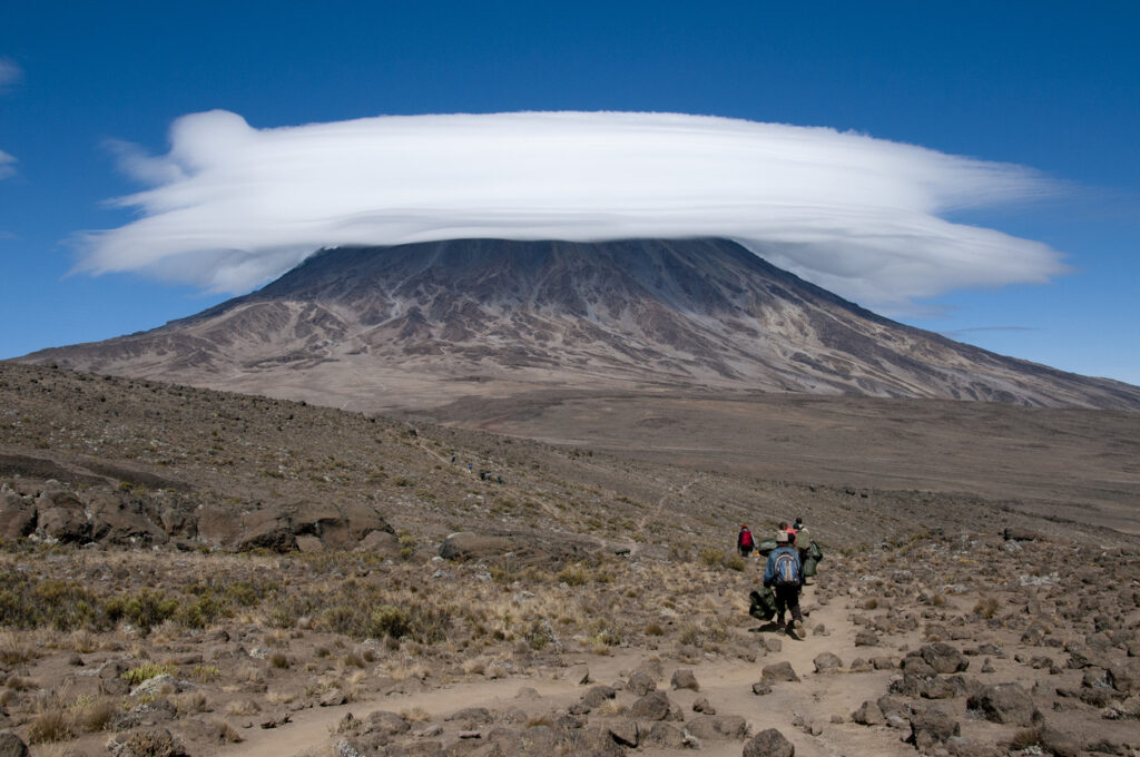 Trekker gehen über karges Hochland auf den Kilimandscharo, dessen Gipfel von einer linsenförmigen Wolke bedeckt ist.