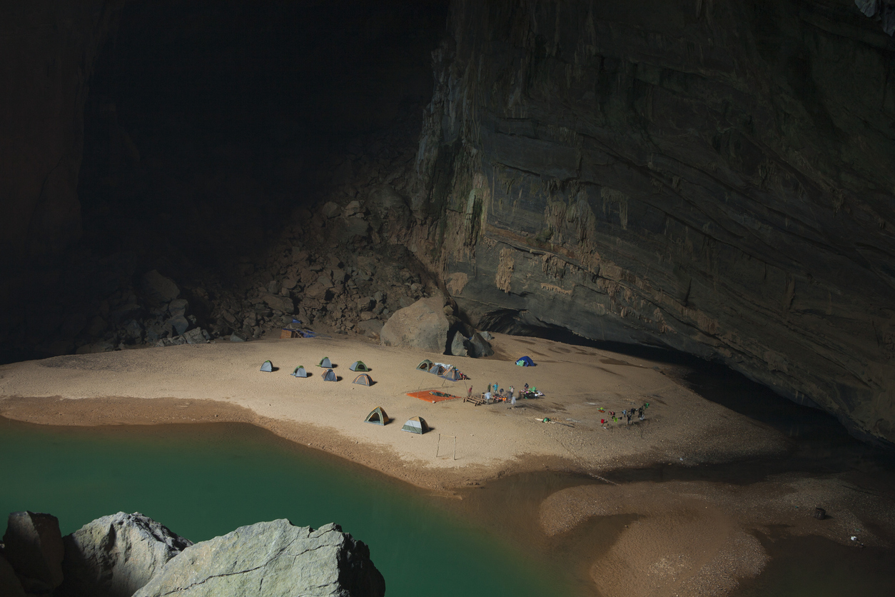 Zelte auf einer sandigen Höhlenbank neben smaragdgrünem Wasser.