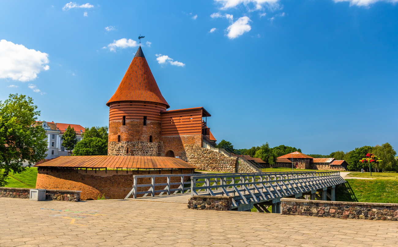 Der runde Backsteinturm der Burg Kaunas steht neben einer Holzbrücke unter blauem Himmel.