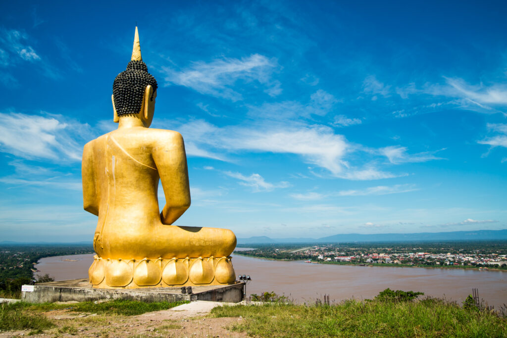 Große goldene Buddha-Statue von hinten über dem Mekong mit Stadtblick und blauem Himmel.