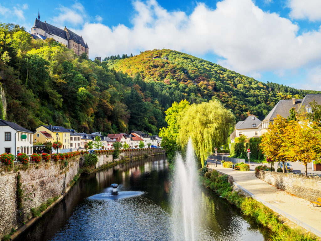 Bunter Häuserreihe am Fluss mit Springbrunnen und Burg auf dem Hügel.