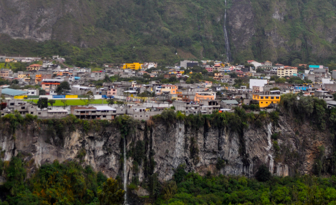 Häuser von Baños de Agua Santa liegen oberhalb einer steilen Felswand mit Wasserfall.
