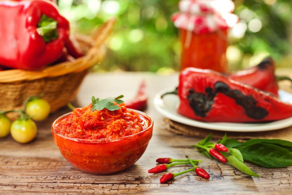 Eine Schale Ajvar mit Petersilie steht auf einem Holztisch, hinten liegen geröstete rote Paprika und kleine Chilis.