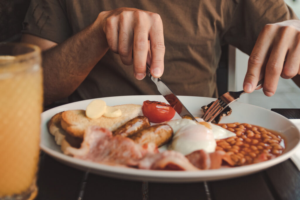 Ein Teller mit Full English Breakfast mit Eiern, Würstchen, Bohnen, Speck, Toast und gebratener Tomate, während eine Person mit Messer und Gabel isst.