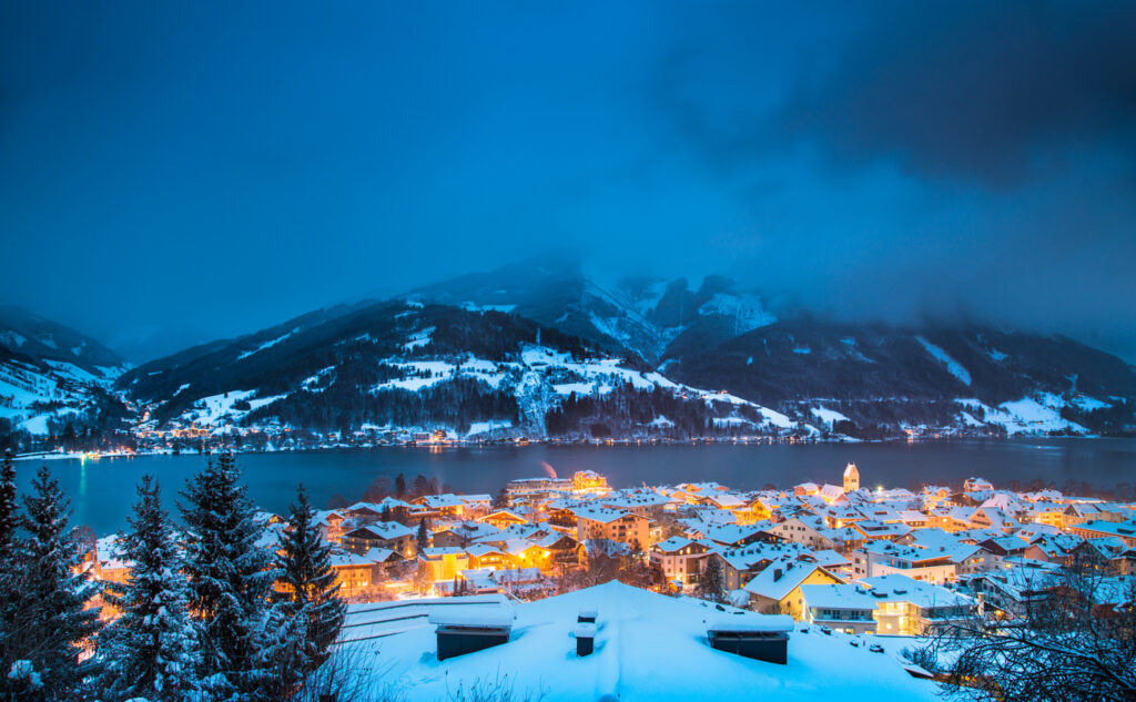 Die Stadt Zell am See liegt verschneit am See, beleuchtet von warmen Lichtern im Abendblau.