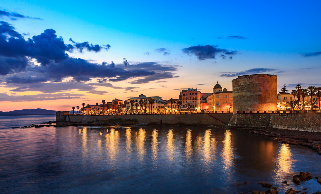 Abendstimmung in Alghero mit beleuchteter Uferpromenade, Stadtmauer und Spiegelungen der Lichter im Meer.