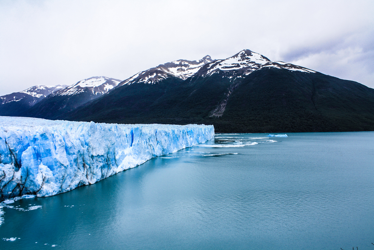 Mächtiger Perito-Moreno-Gletscher ragt als blaue Eiswand in einen türkisfarbenen See vor dunklen Bergen mit Schneeresten.