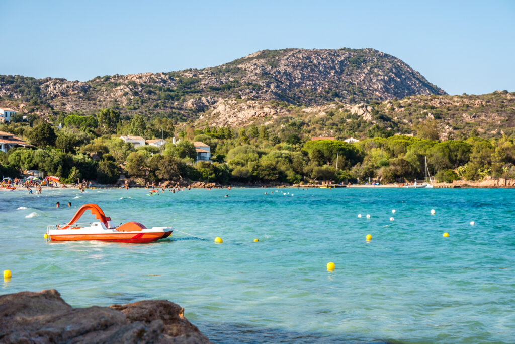 Blick auf den Strand von Porto Istana bei Olbia mit türkisblauem Wasser, vielen Badenden und einem Tretboot im Vordergrund.