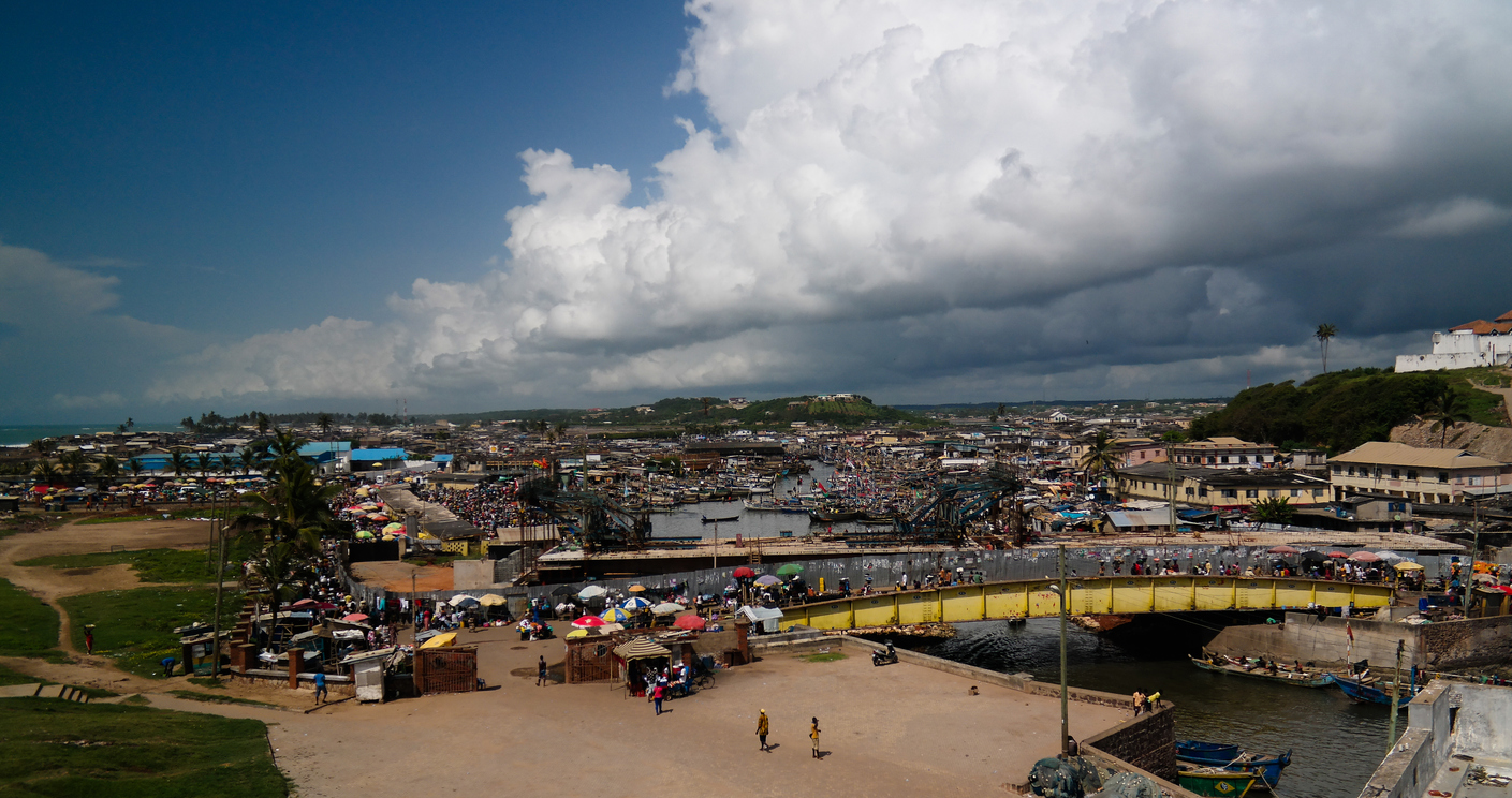 Weitwinkelaufnahme des geschäftigen Hafens und Marktes von Cape Coast & Elmina mit dunklen Wolken am Himmel.