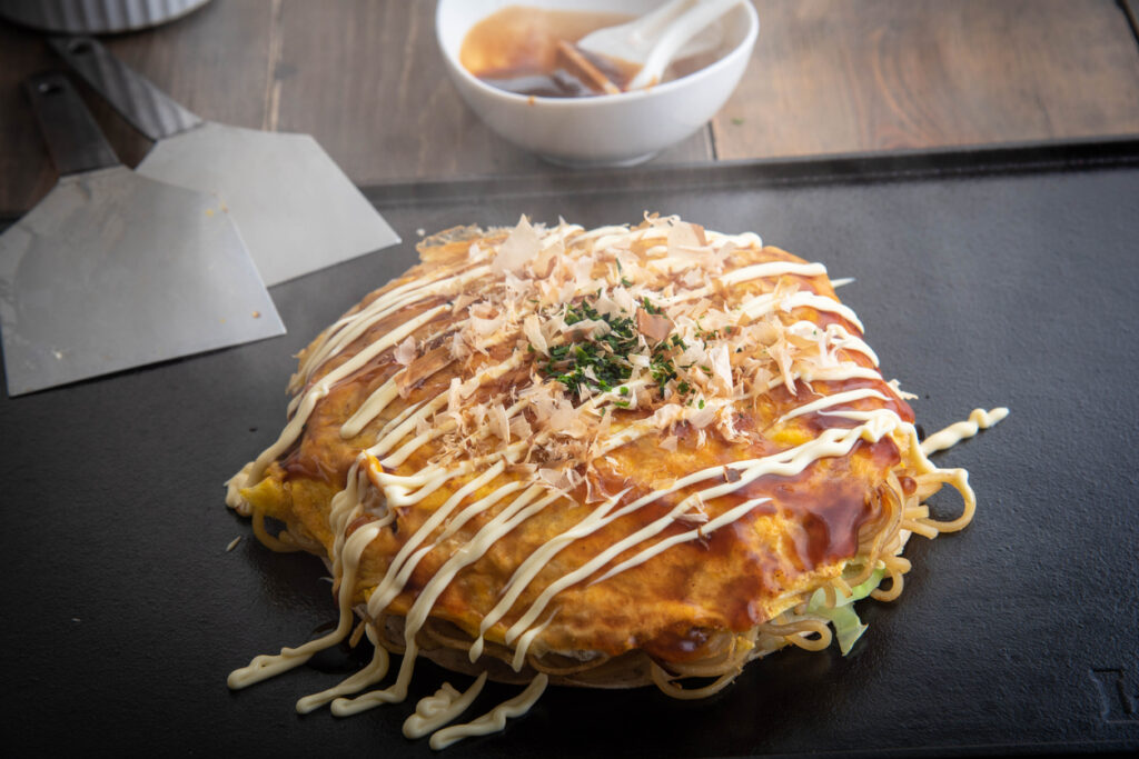 Japanisches Okonomiyaki mit Sauce, Mayonnaise und Bonitoflocken liegt dampfend auf einer Grillplatte.