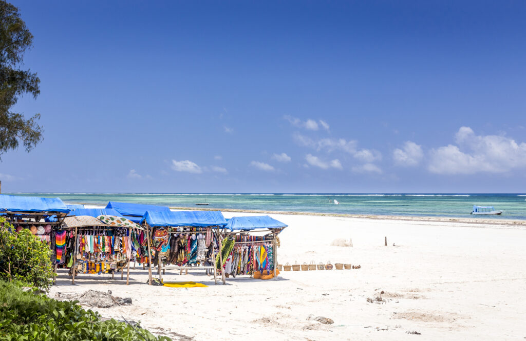 Kleine Verkaufsstände mit bunten Tüchern stehen direkt am weißen Strand von Diani Beach in Kenia.