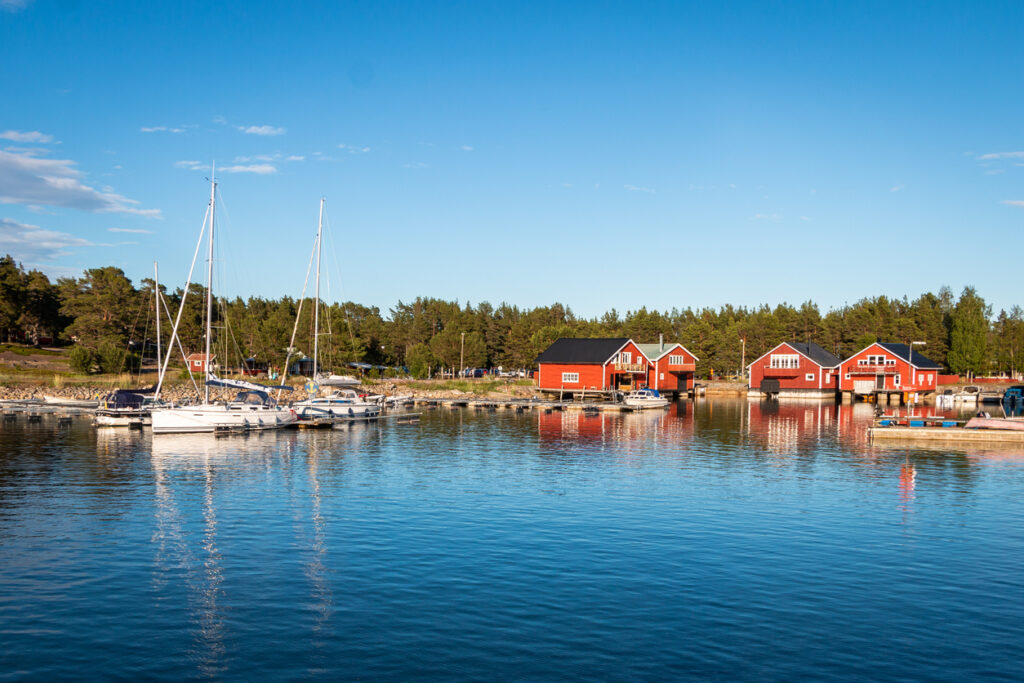 Segelboote liegen im ruhigen Hafenwasser vor roten Holzhäusern am Bottnischen Meerbusen.