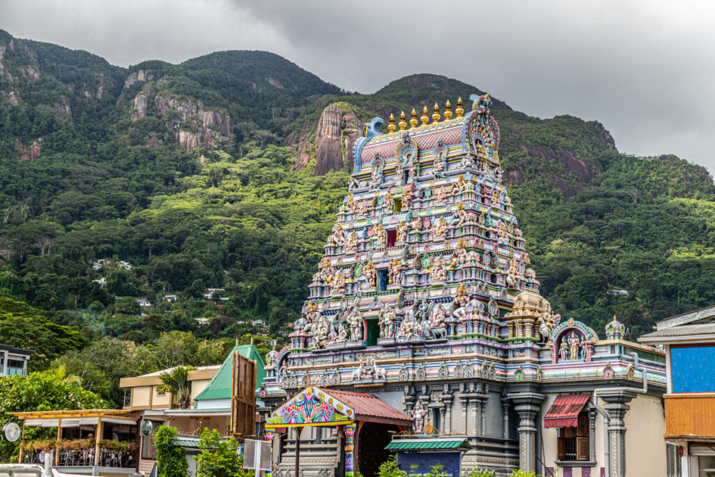 Farbenreicher Hindu Tempel in Victoria auf Mahé, im Hintergrund steigen grüne Berge unter grauem Himmel auf.