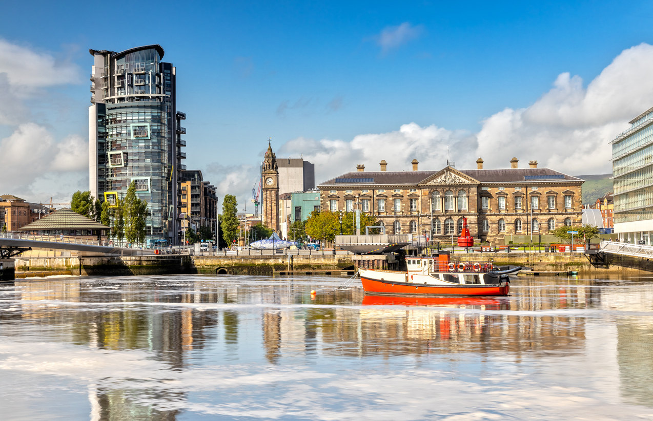 Blick über den Fluss in Belfast mit moderner Architektur, historischen Gebäuden und einem Boot im Vordergrund.