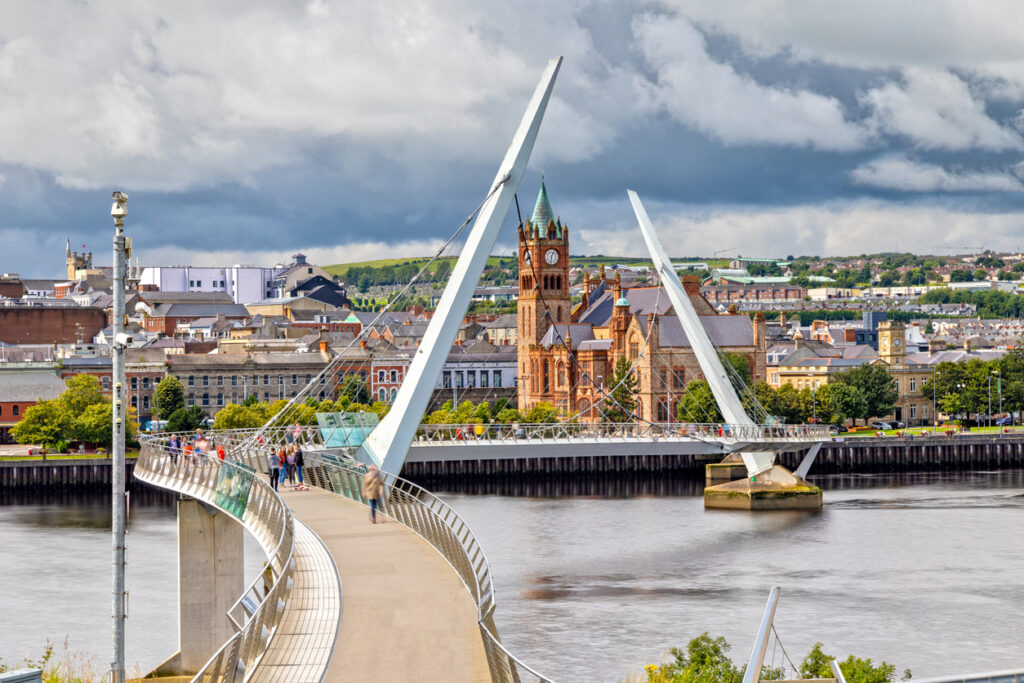 Geschwungene Fußgängerbrücke über dem Fluss in Derry mit Stadtblick und rotem Backsteinbau im Hintergrund.