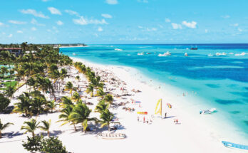 Ein langer Sandstrand in Punta Cana in der Dominikanischen Republik mit Palmen, Badegästen und Booten im türkisblauen Meer.