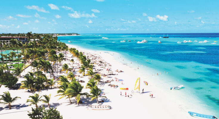 Ein langer Sandstrand in Punta Cana in der Dominikanischen Republik mit Palmen, Badegästen und Booten im türkisblauen Meer.