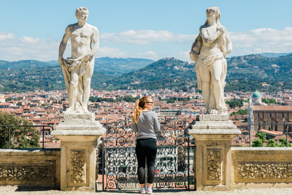 Eine Frau blickt von einer Terrasse mit Statuen auf die Dächer von Florenz.