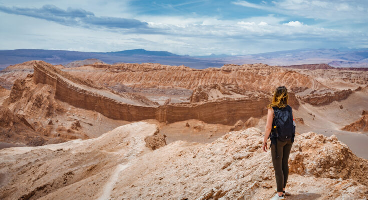Eine Wanderin steht auf einem Felsplateau bei San Pedro de Atacama, Chile, und blickt über die Wüstenlandschaft.