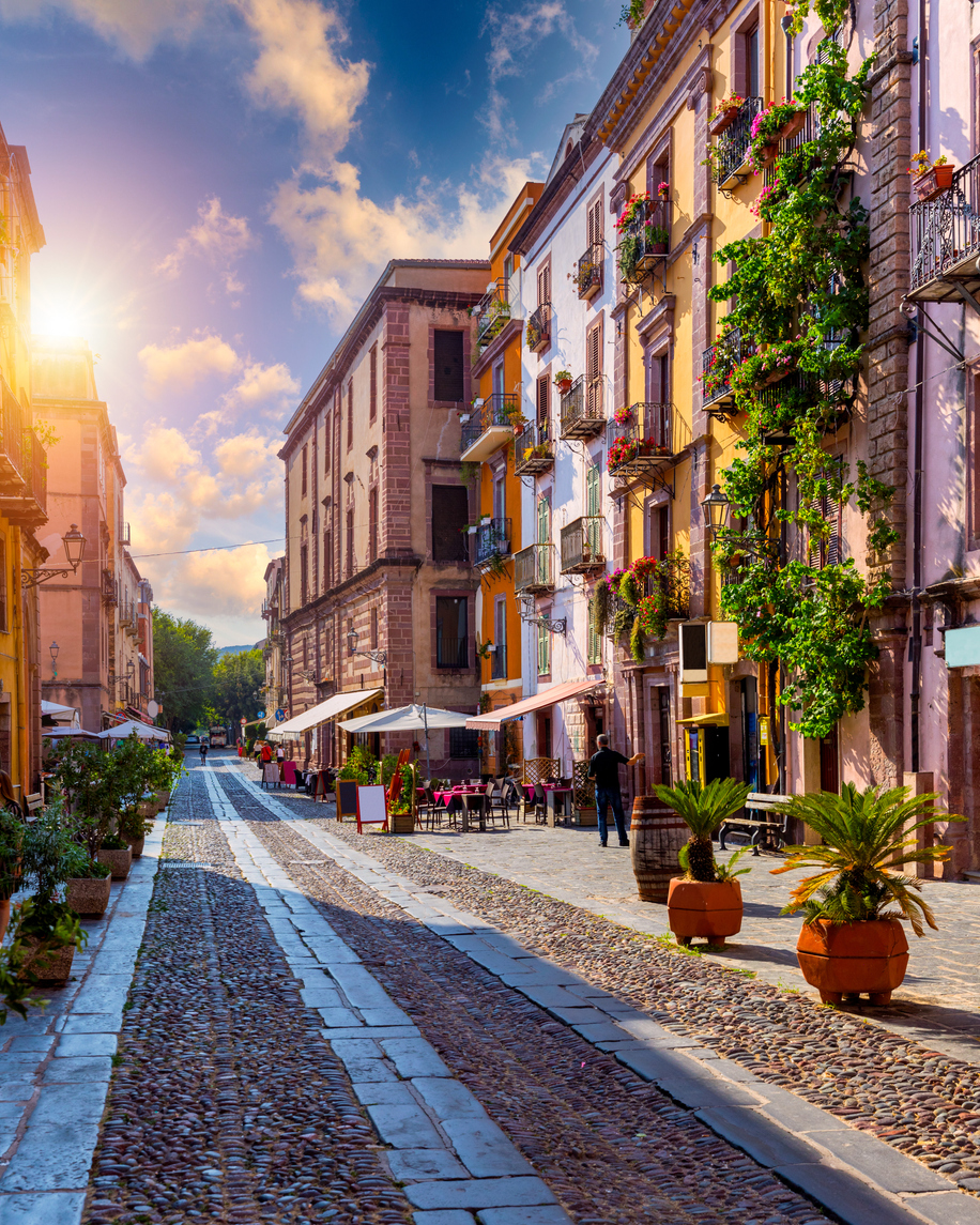 Sonnenbeschienene Altstadtgasse mit bunten Häusern, Cafés und Kopfsteinpflaster in Bosa auf Sardinien.