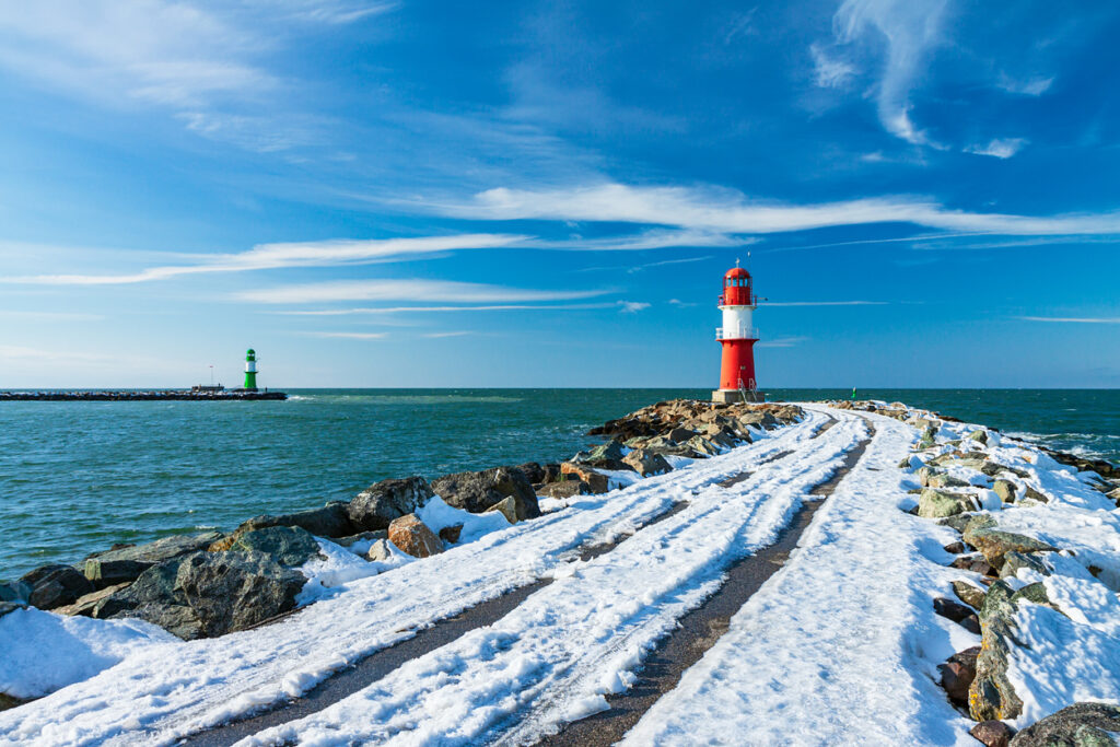 Roter Leuchtturm am Ende einer verschneiten Mole in Warnemünde, im Hintergrund ein grüner Leuchtturm und die winterliche Ostsee.