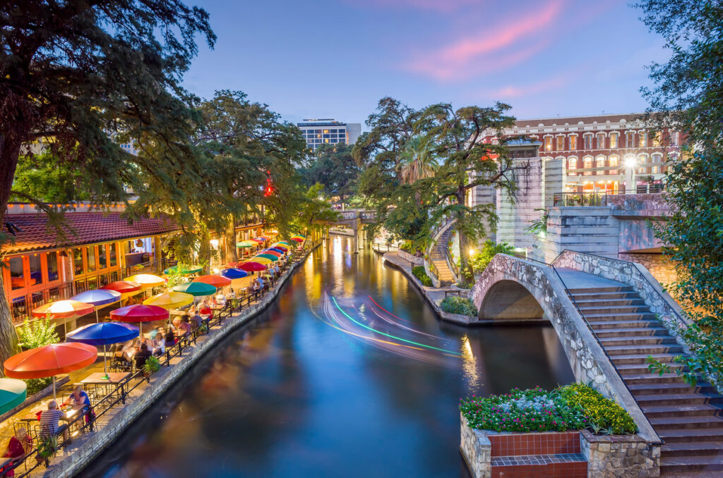 Abendstimmung am River Walk in San Antonio, Texas, mit bunten Sonnenschirmen, Restaurants und beleuchteten Booten auf dem Wasser.