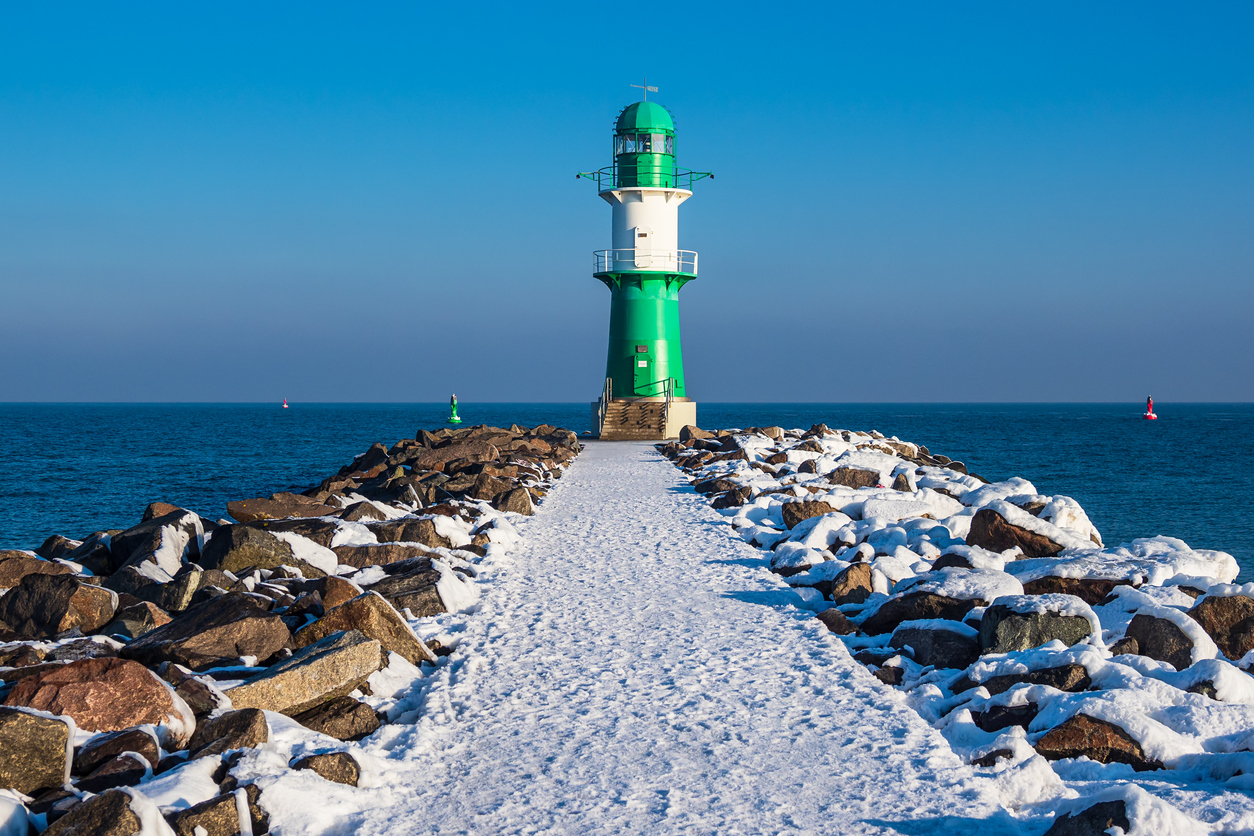 Grüner Leuchtturm am Ende einer schneebedeckten Steinmole in Warnemünde, umgeben von ruhiger blauer Ostsee.