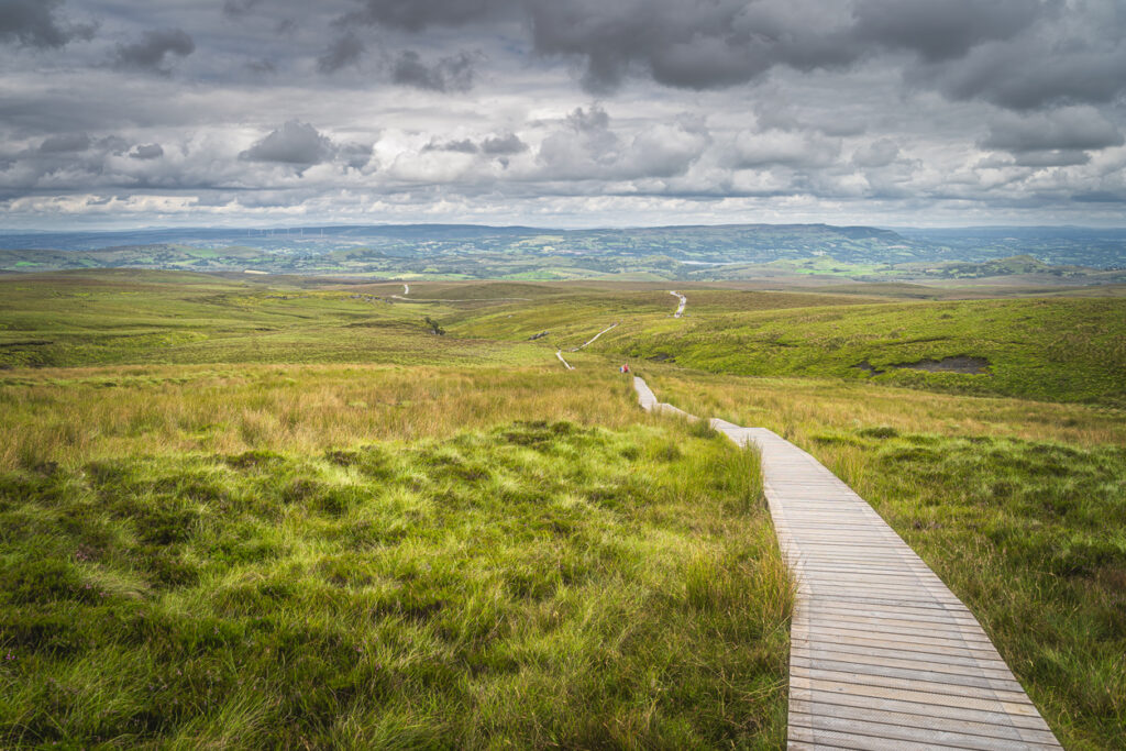 Langer Holzsteg führt durch Moorlandschaft im Cuilcagh Mountain Park in Nordirland unter dramatischen Wolken.