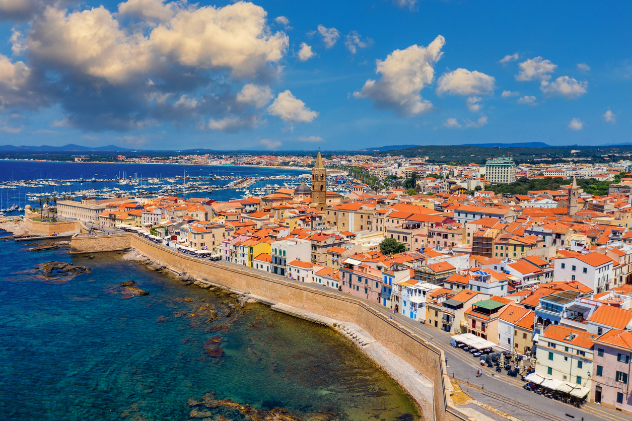 Blick auf die Altstadt von Alghero mit Stadtmauer, orangefarbenen Dächern und tiefblauem Meer an der Nordwestküste Sardiniens.