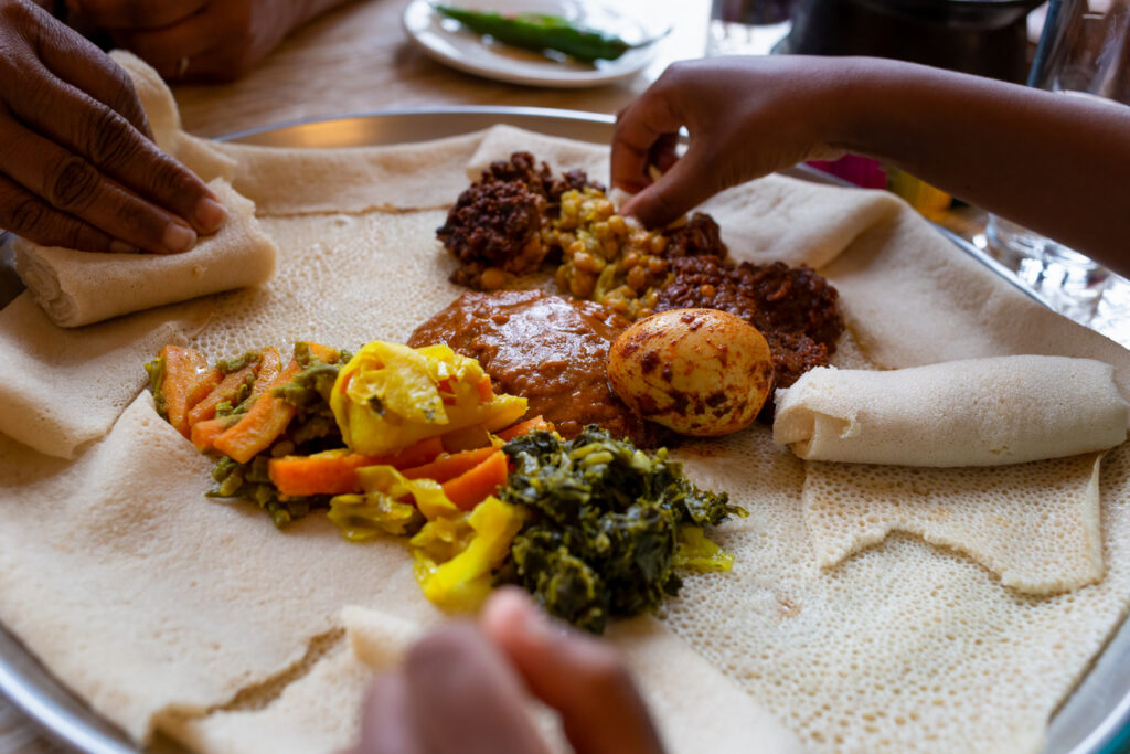 Hände greifen zu einem großen Teller mit Vegetarisches Injera-Menü mit Shiro, Linsen und Ei, dazu Gemüse und Soßen auf weichem Fladenbrot.