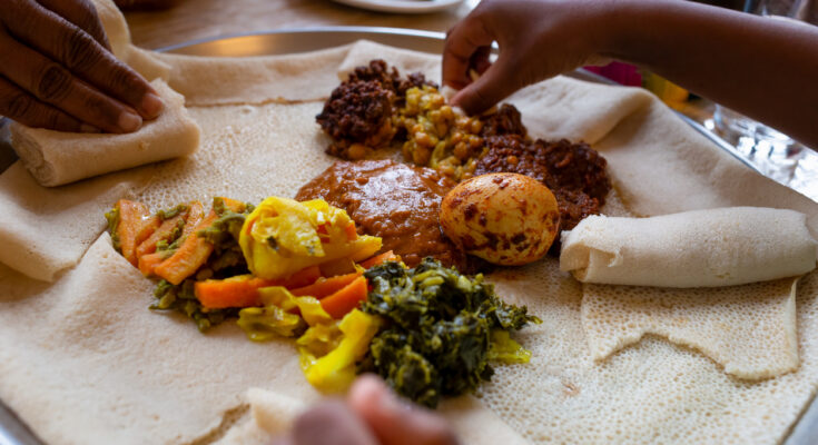 Hände greifen zu einem großen Teller mit Vegetarisches Injera-Menü mit Shiro, Linsen und Ei, dazu Gemüse und Soßen auf weichem Fladenbrot.