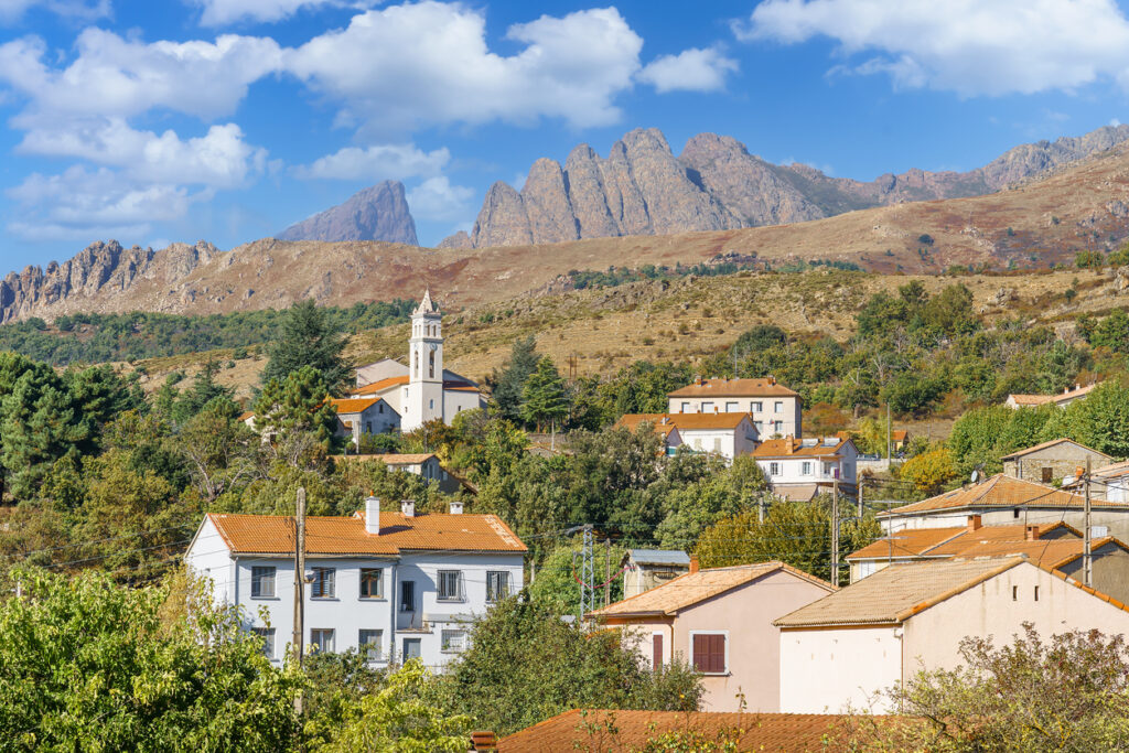In Calacuccia stehen helle Häuser und eine Kirche mit Glockenturm vor einer wilden Bergkulisse auf Korsika.
