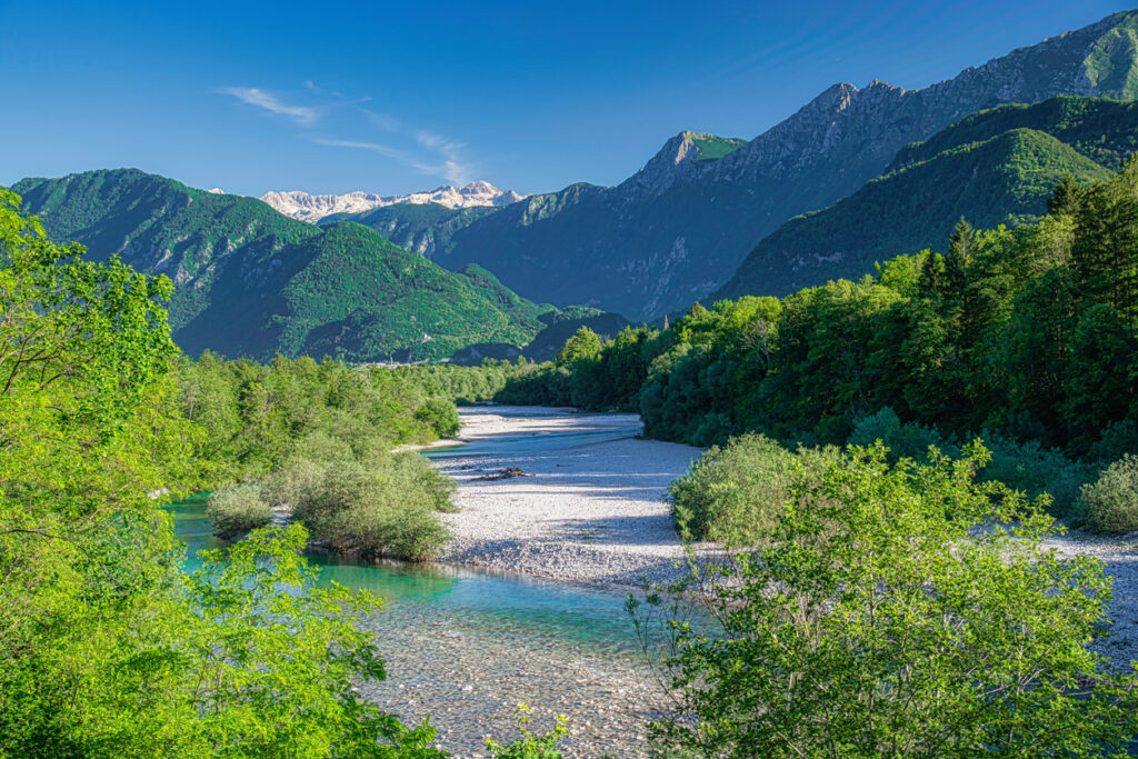 Panoramablick auf das Soča-Tal mit türkisblauem Flussbett, grünen Wäldern und hohen Bergen im Hintergrund.