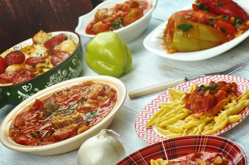 Verschiedene ungarische Gerichte stehen in Schalen und auf Tellern auf einem hellen Holztisch, darunter gefüllte Paprika, Eintöpfe und Nudeln.