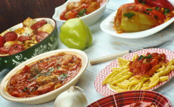 Verschiedene ungarische Gerichte stehen in Schalen und auf Tellern auf einem hellen Holztisch, darunter gefüllte Paprika, Eintöpfe und Nudeln.