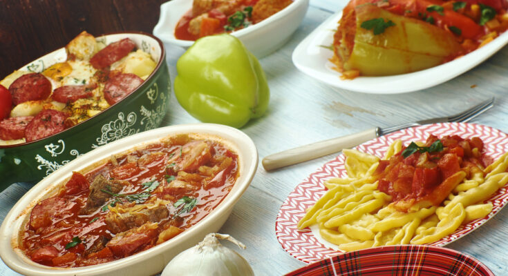 Verschiedene ungarische Gerichte stehen in Schalen und auf Tellern auf einem hellen Holztisch, darunter gefüllte Paprika, Eintöpfe und Nudeln.