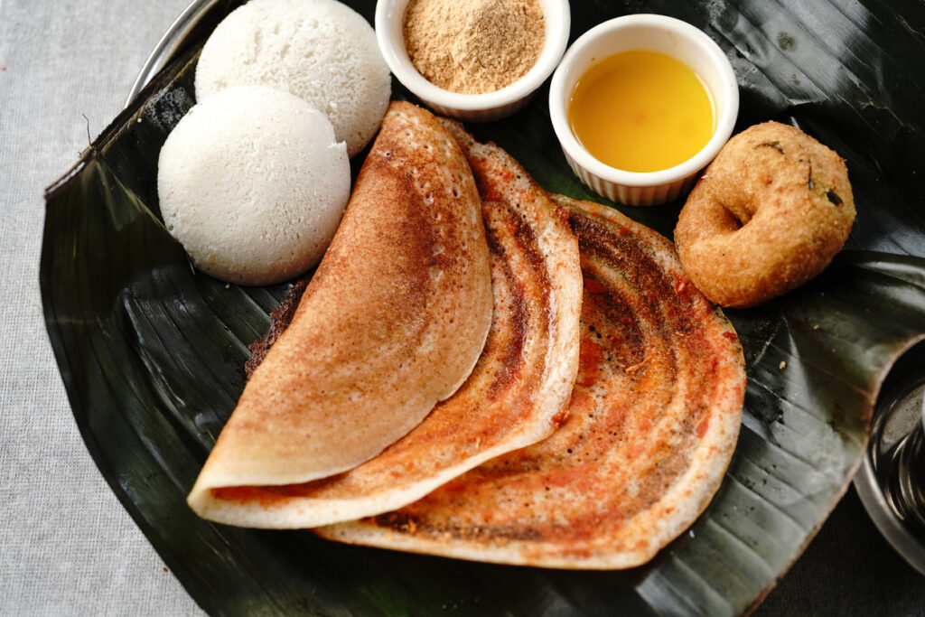 Dosas liegen auf einem Bananenblatt mit Idli, Vada und kleinen Schalen mit Chutney und Ghee.