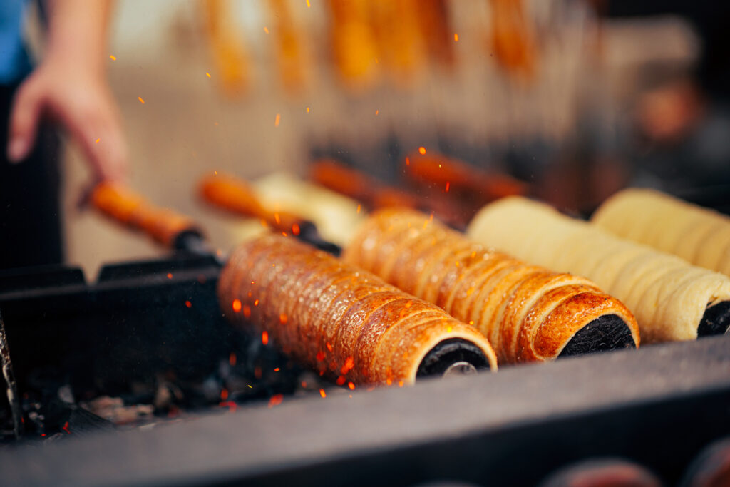 Mehrere Kürtőskalács-Rollen drehen sich auf Metallspießen über einem Grill mit sprühenden Funken.