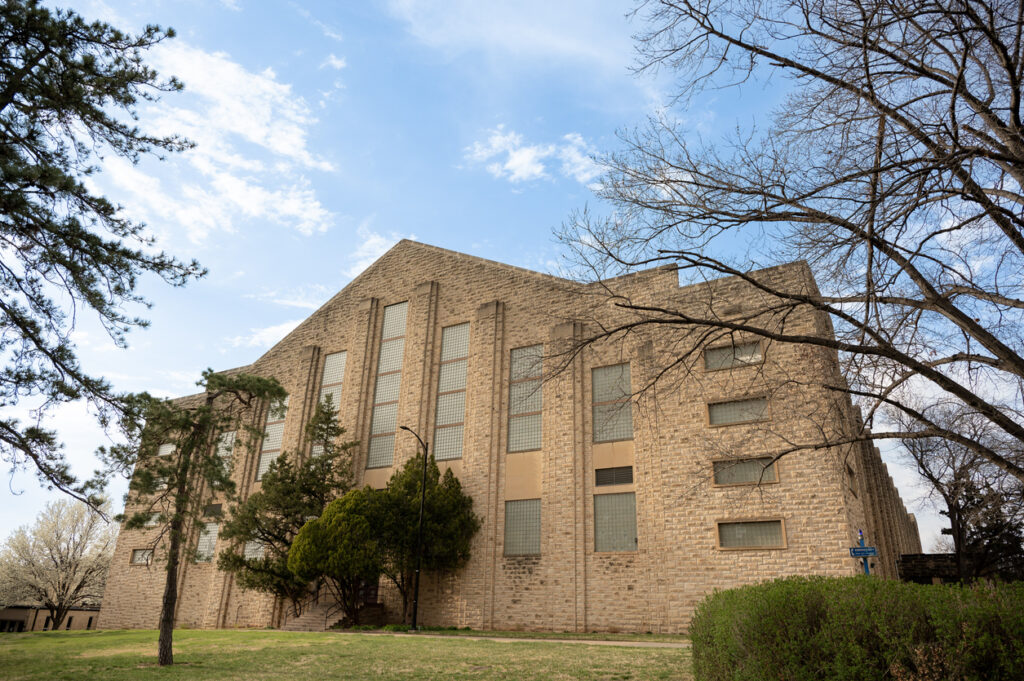 Sandstein-Gebäude der Kansas State University mit hohen Fenstern, umrahmt von Bäumen unter blauem Himmel.