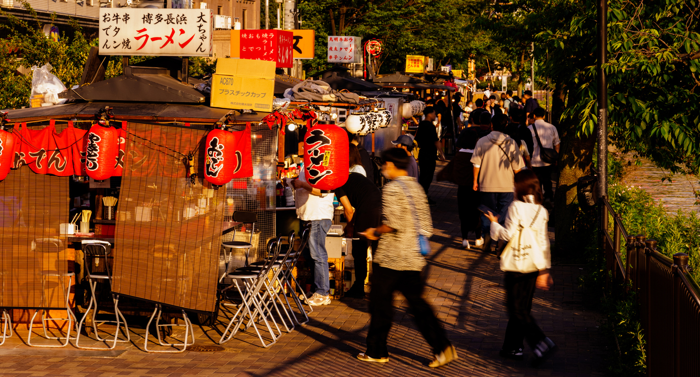 Menschen schlendern an beleuchteten Streetfood-Ständen mit roten Laternen in Fukuoka vorbei.
