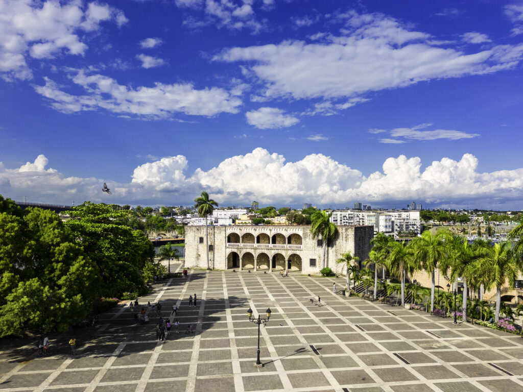 Ein großer Platz mit Palmen führt in Santo Domingo auf ein historisches Steingebäude mit Arkaden zu.