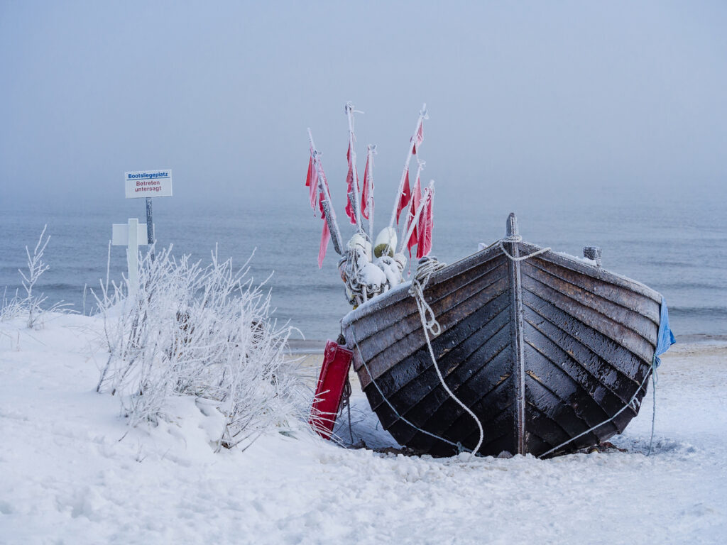 Ein eingefrorenes Fischerboot im Winter an der Ostsee steht im Schnee vor nebligem Meer.