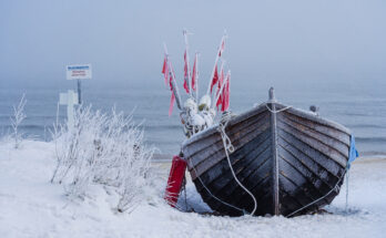Ein eingefrorenes Fischerboot im Winter an der Ostsee steht im Schnee vor nebligem Meer.