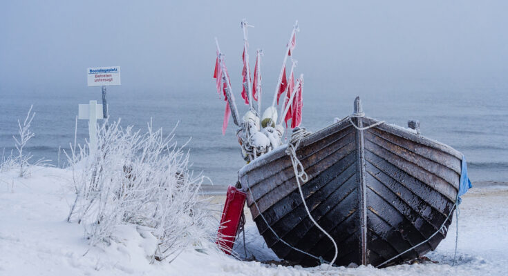 Ein eingefrorenes Fischerboot im Winter an der Ostsee steht im Schnee vor nebligem Meer.